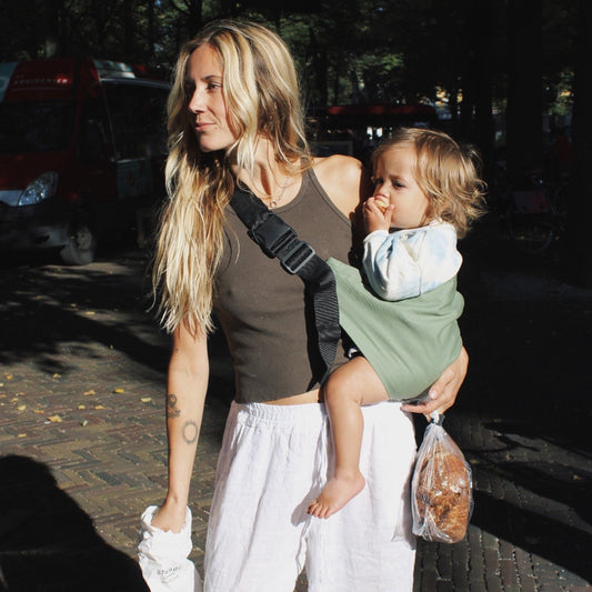 Woman walking outdoors with a child in a sling and a bag of bread.