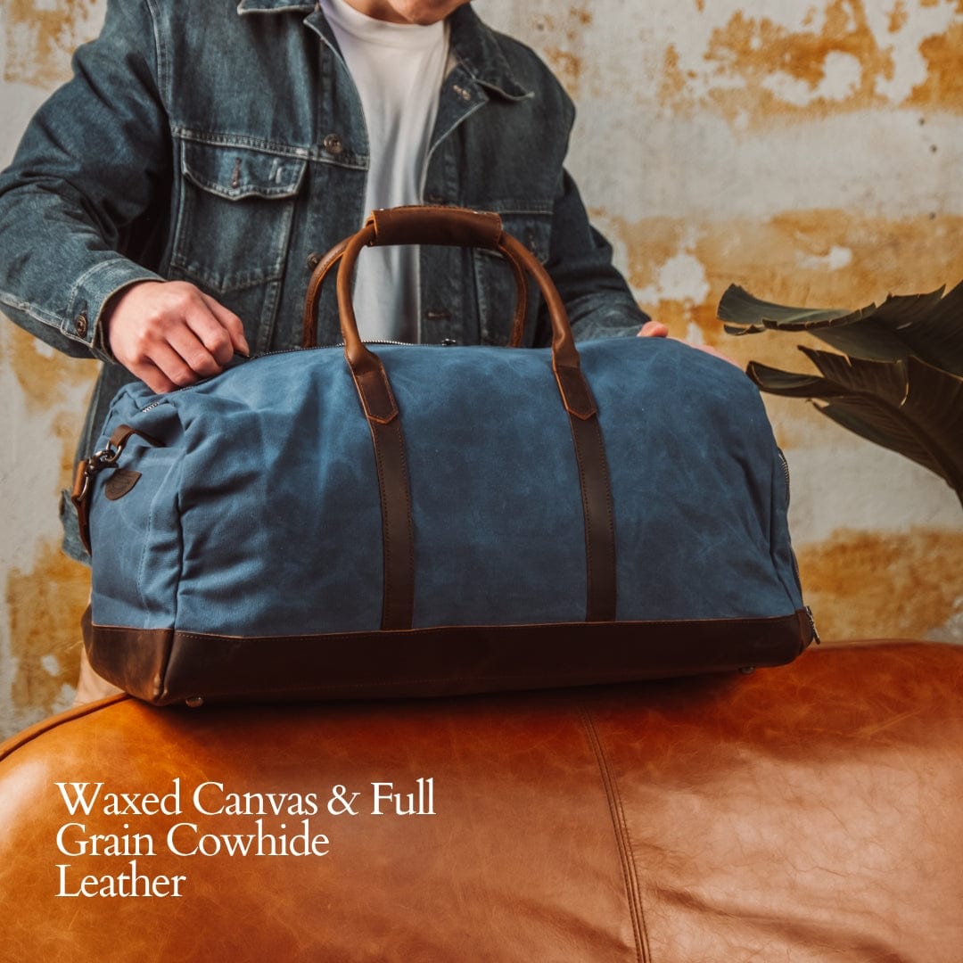 Man holding a blue waxed canvas duffel bag with brown leather handles against a textured wall.