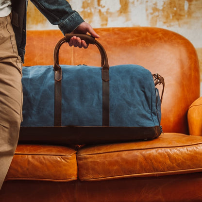 Man holding a blue duffel bag with brown handles and straps on an orange leather couch.