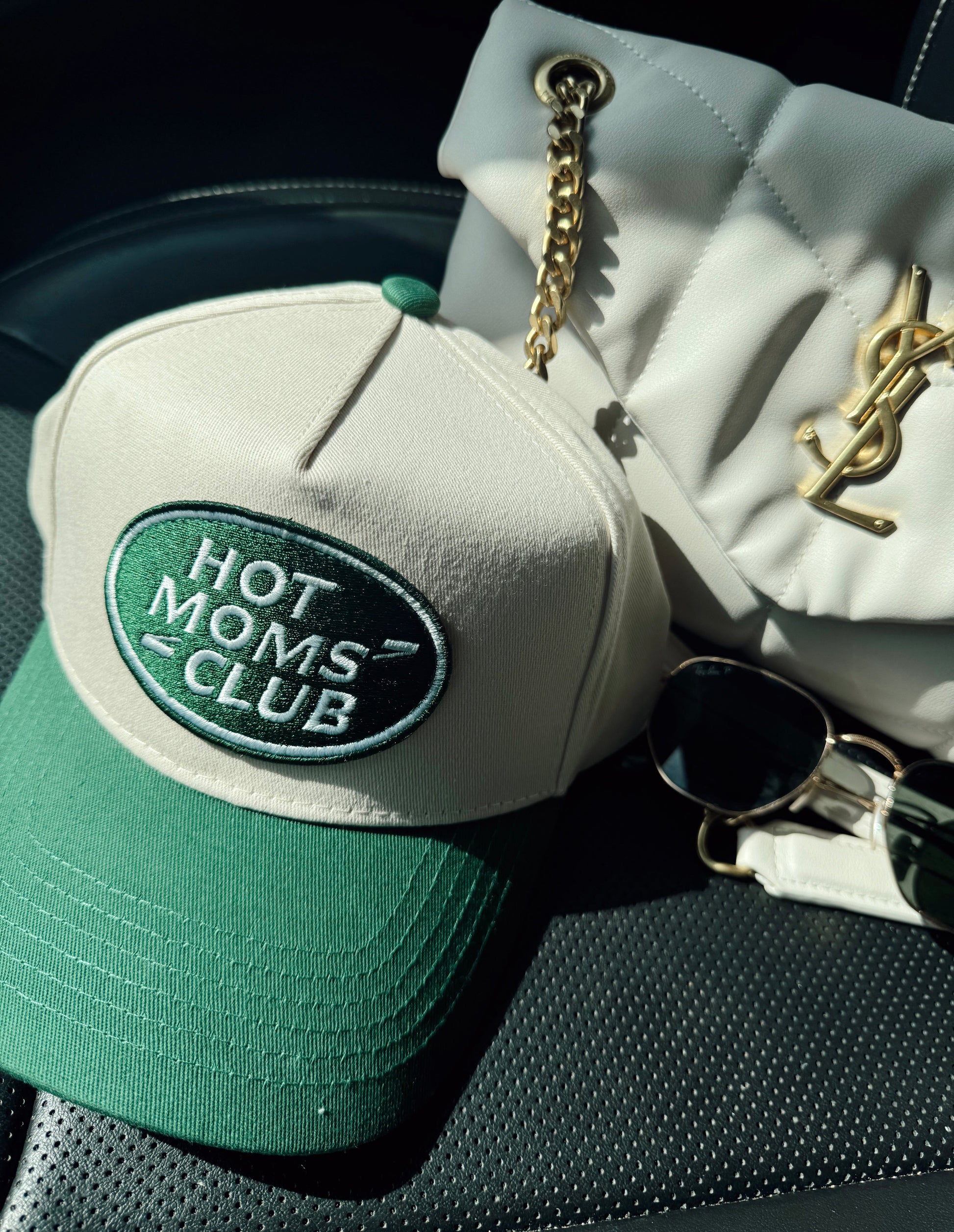 Green cap with 'Hot Moms Club' logo on a car seat next to a YSL handbag.