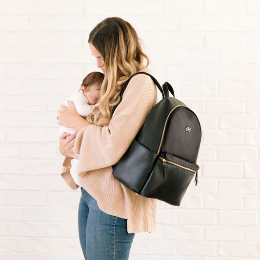Mom holding her baby with the Freshly Picked Ebony black vegan leather diaper backpack with matte gold hardware on her back