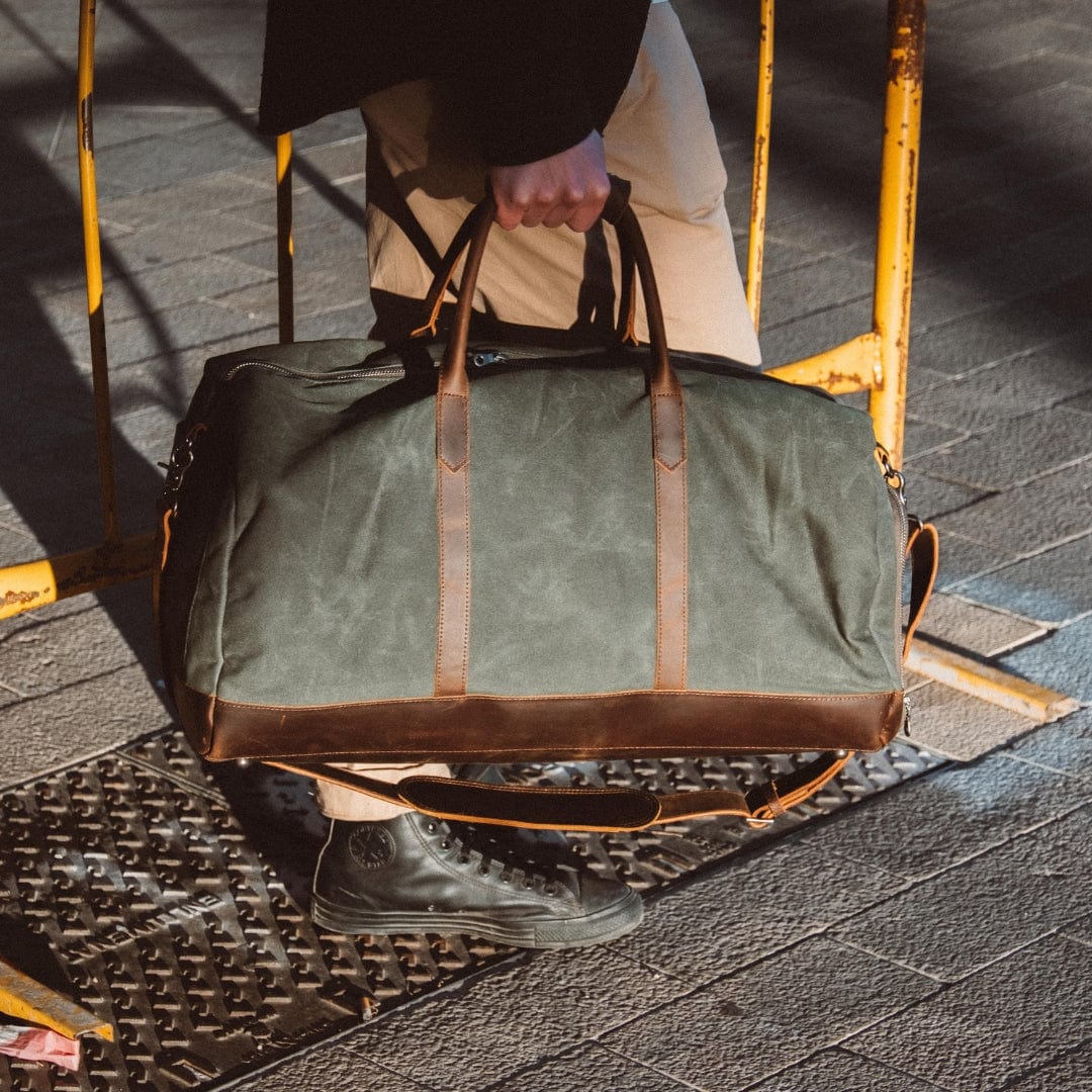 Man holding a green duffel bag with brown leather straps on a sidewalk.