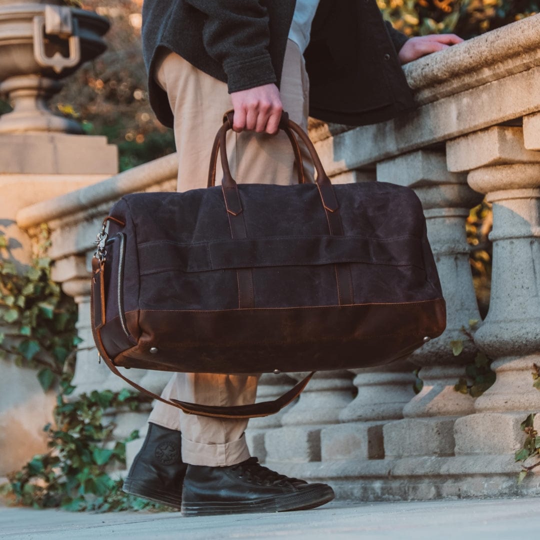Man holding a dark brown leather duffel bag outdoors.