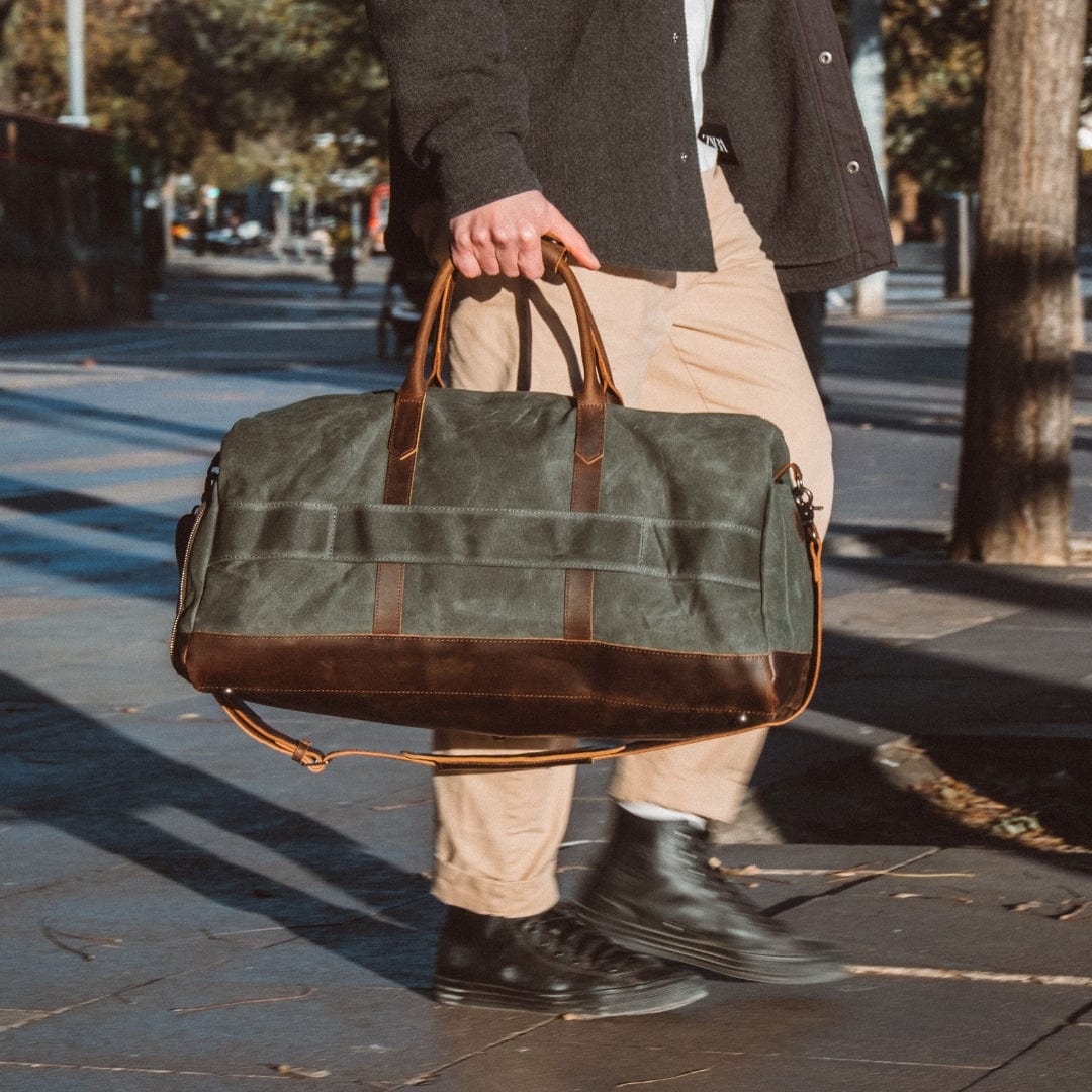 Manholding a green duffel bag with brown leather straps on a city street.
