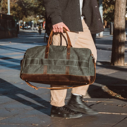 Manholding a green duffel bag with brown leather straps on a city street.