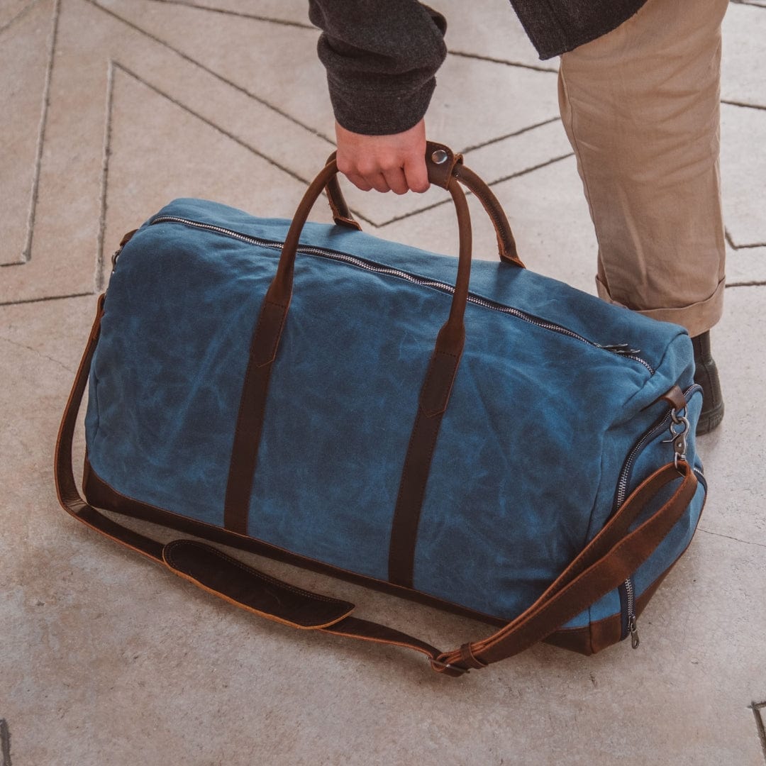 Blue duffel bag with brown straps held by a person on a stone pavement.
