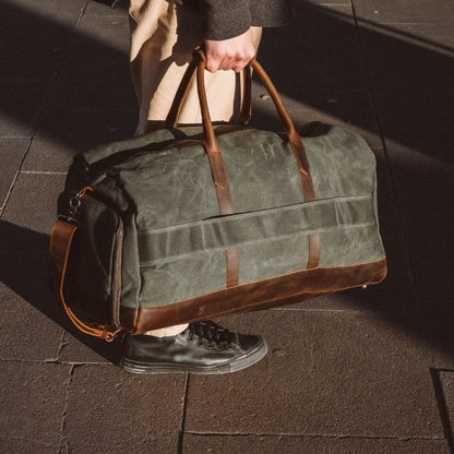 Man holding a green duffel bag with brown leather straps on a tiled floor.
