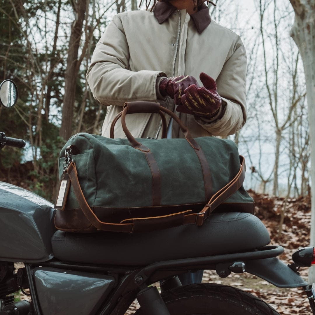 Person sitting on a motorcycle with a green duffel bag on their lap, surrounded by trees.