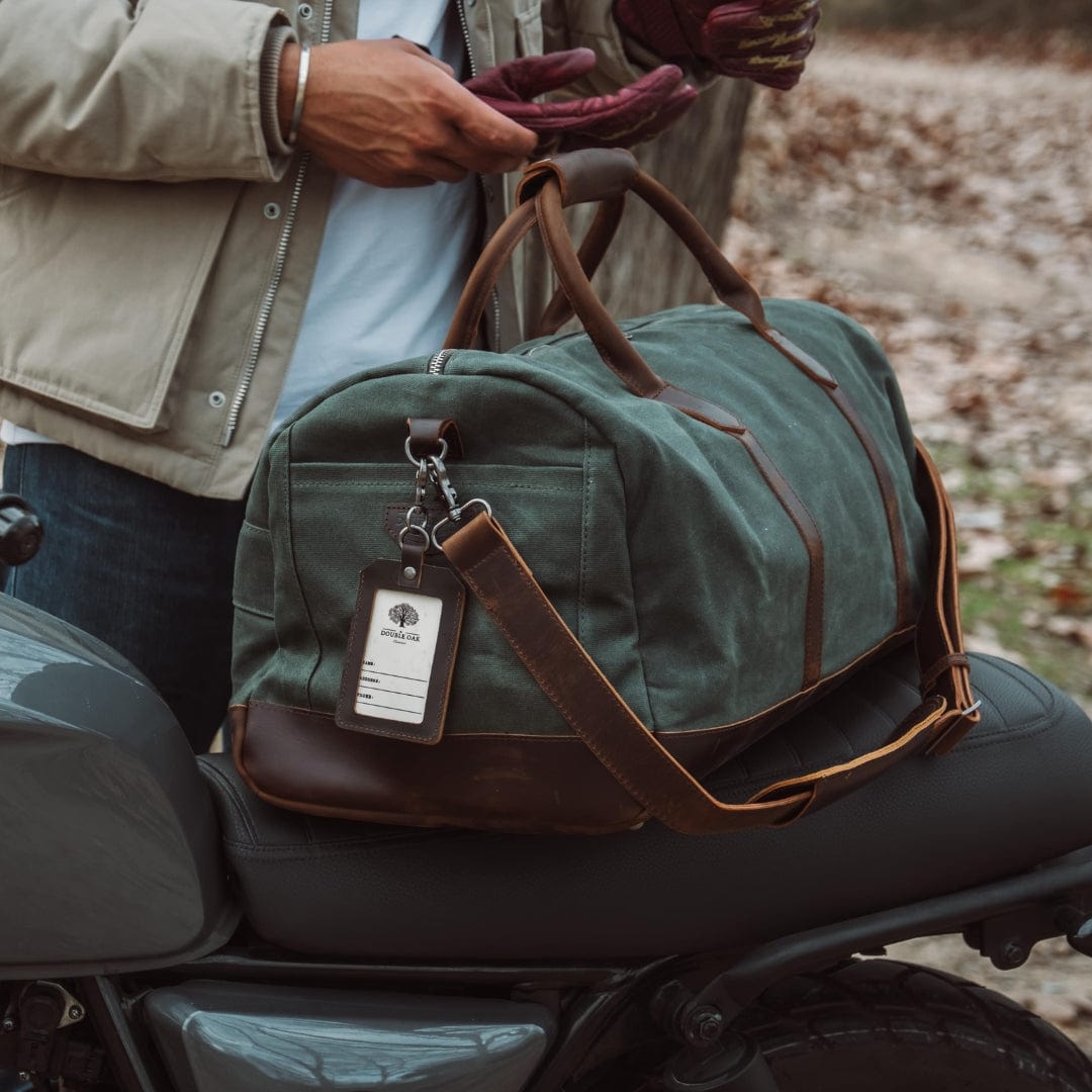 Green duffel bag with brown leather straps on a motorcycle seat, person holding it.