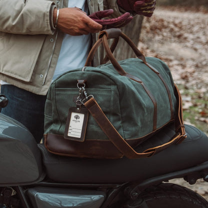 Green duffel bag with brown leather straps on a motorcycle seat, person holding it.