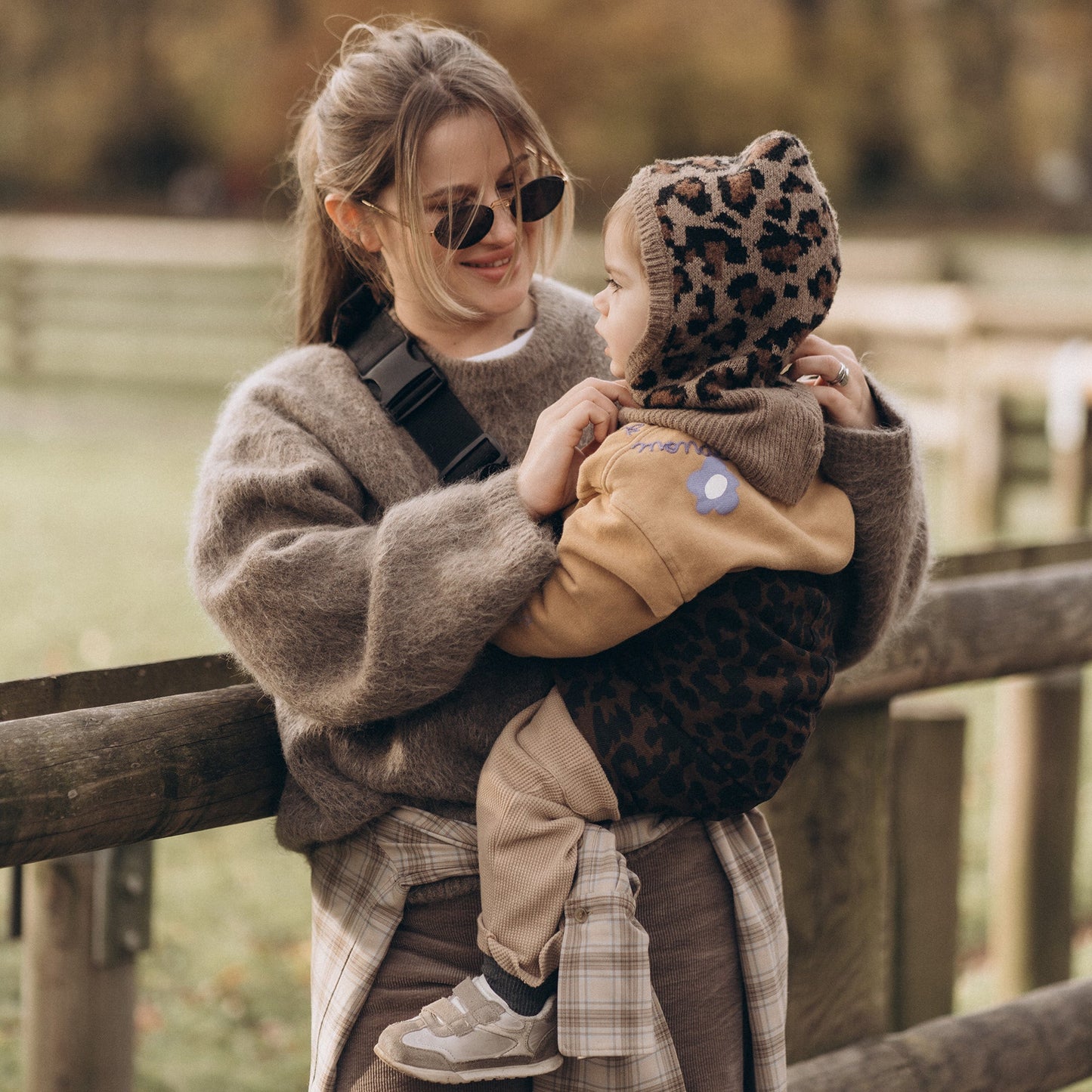Woman holding a child wearing a leopard print hat by a wooden fence