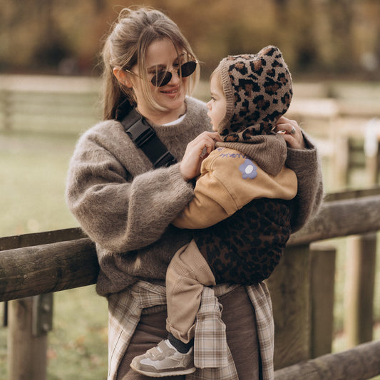 Woman holding a child wearing a leopard print hat by a wooden fence